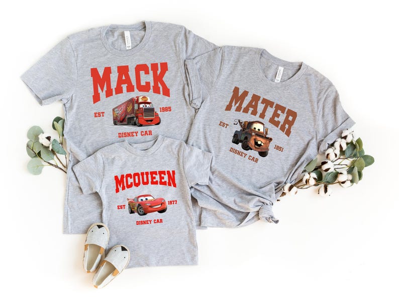 May include: Three gray t-shirts with Cars characters and text. The largest shirt says "MACK EST 1985 DISNEY CAR" with a red truck. The medium shirt says "MATER EST 1951 DISNEY CAR" with a brown tow truck. The smallest shirt says "MCQUEEN EST 1977 DISNEY CAR" with a red race car.
