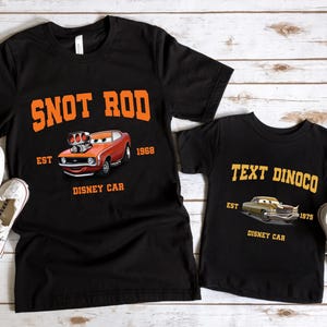 May include: Two black t-shirts with a Cars movie theme. The larger shirt features an orange car with the text "Snot Rod Est 1968 Disney Car". The smaller shirt features a gold car with the text "Text Dinoco Est 1975 Disney Car".