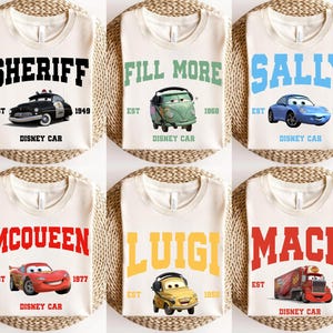 May include: Six white t-shirts with different Disney Cars characters and names printed on them. The shirts feature the characters Sheriff, Fill More, Sally, McQueen, Luigi, and Mack. Each shirt has the text "Disney Car" and the year the character was introduced.
