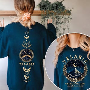 May include: A teal blue sweatshirt with a gold graphic design featuring a mountain range, crescent moon, and stars. The text "VELARIS CITY OF STARLIGHT" is printed on the sweatshirt. The text "To the stars who listen and the dreams that are answered" is printed below the graphic.