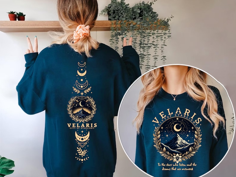 May include: A teal blue sweatshirt with a gold graphic design featuring a mountain range, crescent moon, and stars. The text "VELARIS CITY OF STARLIGHT" is printed on the sweatshirt. The text "To the stars who listen and the dreams that are answered" is printed below the graphic.