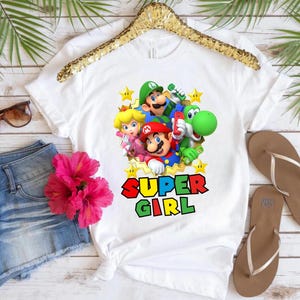 May include: White t-shirt with a graphic print of Mario, Luigi, Peach, and Yoshi characters from the Super Mario video game series. The text "SUPER GIRL" is printed below the characters.