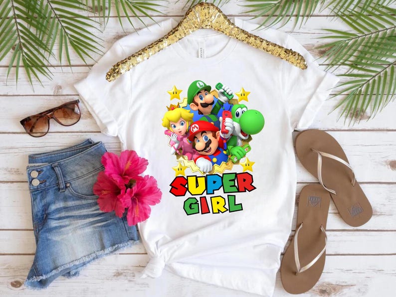 May include: White t-shirt with a graphic print of Mario, Luigi, Peach, and Yoshi characters from the Super Mario video game series. The text "SUPER GIRL" is printed below the characters.