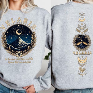 May include: Grey sweatshirt with a graphic design featuring a mountain range, crescent moon, and stars. The text "VELARIS CITY OF STARLIGHT" is printed on the front of the sweatshirt. The back of the sweatshirt features a vertical design with a mountain range, crescent moon, and stars.