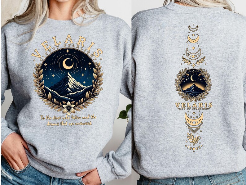 May include: Grey sweatshirt with a graphic design featuring a mountain range, crescent moon, and stars. The text "VELARIS CITY OF STARLIGHT" is printed on the front of the sweatshirt. The back of the sweatshirt features a vertical design with a mountain range, crescent moon, and stars.