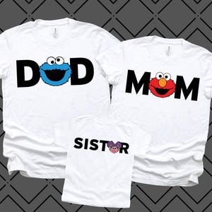 May include: White t-shirts with black text and character graphics. One shirt says "DAD" with a blue Cookie Monster graphic. Another says "MOM" with an Elmo graphic. A smaller shirt says "SISTER" with a purple character graphic.