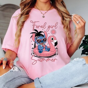 以下が含まれることがあります： ピンク色のTシャツ。リロ＆スティッチのスティッチがサングラスをかけ、フラミンゴの浮き輪に乗って飲み物を持っているグラフィック。テキスト「Feral girl Summer」がグラフィックの上と下にあります。