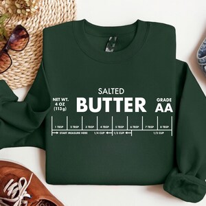 May include: Dark green sweatshirt with the words "SALTED BUTTER" in large white letters. Below the text is a measurement guide. The sweatshirt also has the text "NET WT. 4 OZ (113g)" and "GRADE AA".