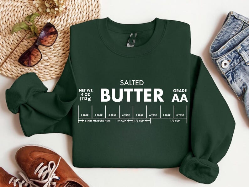 May include: Dark green sweatshirt with the words "SALTED BUTTER" in large white letters. Below the text is a measurement guide. The sweatshirt also has the text "NET WT. 4 OZ (113g)" and "GRADE AA".