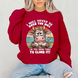 Puede incluir: Sudadera roja con una vaca de dibujos animados y el texto "I WILL NEVER BE OVER THE HILL I'M TOO DARN TIRED TO CLIMB IT!"