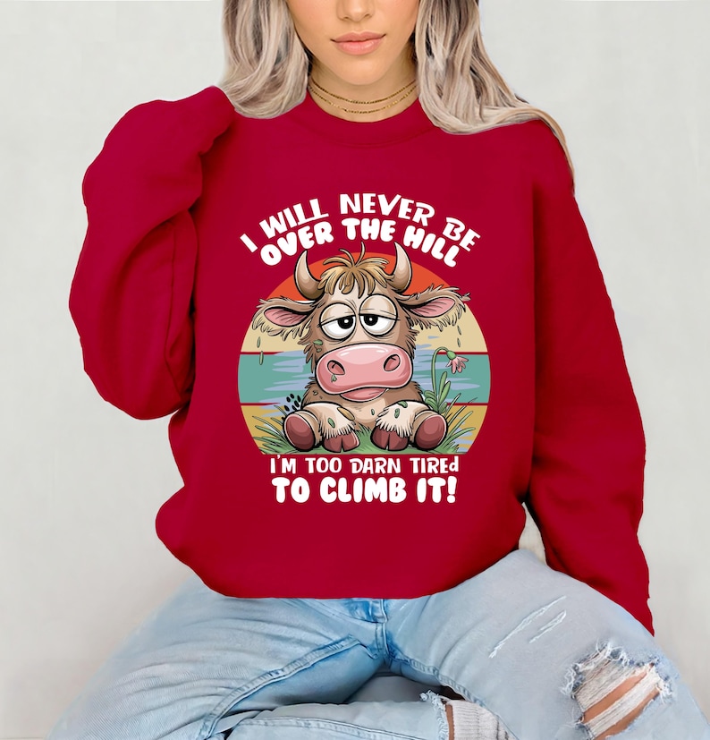 Puede incluir: Sudadera roja con una vaca de dibujos animados y el texto "I WILL NEVER BE OVER THE HILL I'M TOO DARN TIRED TO CLIMB IT!"