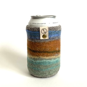 May include: A felted wool can cooler sleeve featuring horizontal stripes in shades of grey, blue, brown, and teal. A rectangular leather tag with a brass rivet and a circular logo depicting a shovel and rake is attached to the top edge.