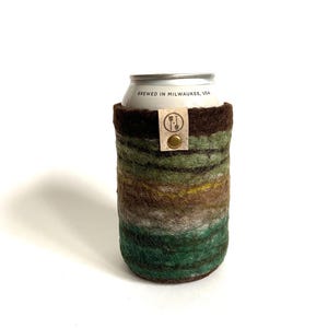 May include: A green, brown, and white striped felt can cooler with a brown leather strap and a brass button. The can cooler says "Brewed in Milwaukee, USA" on the can.