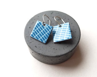 Puede incluir: Un par de pendientes de esmalte azul y blanco con un patrón geométrico. Los pendientes son de forma cuadrada y tienen ganchos de plata.