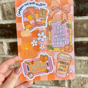 May include: A rectangular sticker with an orange and white checkered background. The sticker features illustrations of a cardigan, pumpkins, flowers, a bow, and a coffee cup. Text includes "Cardigan and book weather", "Cozy up with your Kindle", and "Pumpkin Spice Junkie".