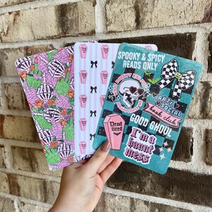 May include: Three decorative rectangular cards with spooky themes. One card features a skeleton hand and roses on a pink and green background. Another has pink coffins and black bows. The third card is teal with a skull and the text "Good Ghoul".