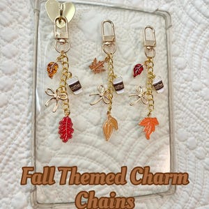 May include: Fall-themed charm chains with gold-colored hardware. Each chain features a coffee cup charm, leaf charms in red, orange, and gold, and a bow charm. The text on the image reads "Fall Themed Charm Chains" and "Adhesive Heart Included".