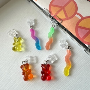 Könnte beinhalten: Sechs bunte Gummibärchen-Anhänger, darunter ein gelber Gummibär, ein orangefarbener Gummibär, ein roter Gummibär, ein grüner Gummibärchenwurm, ein blauer Gummibärchenwurm und ein orangefarbener Gummibärchenwurm. Die Anhänger sind an durchsichtigen Handy-Anhängern aus Kunststoff befestigt.