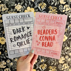 May include: Two decorative guest check cards. One is light gray with the text "DARK ROMANCE GIRLIE". The other is pink with "READERS GONNA READ". Both have a glittery effect and the words "GUEST CHECK" at the top.