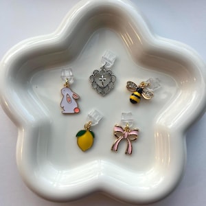 Può includere: Una collezione di cinque ciondoli colorati, tra cui un coniglio bianco, un cuore con una croce, un'ape, un limone e un fiocco rosa. Ogni ciondolo è attaccato a un supporto per orecchini trasparente e presentato su un piatto bianco a forma di nuvola.