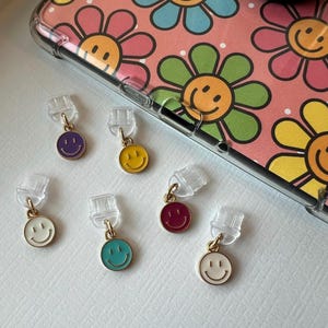 Puede incluir: Seis encantos de caritas sonrientes de colores con detalles dorados. Los encantos están unidos a fundas de teléfono de plástico transparente.