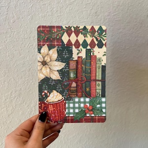 May include: A rectangular Christmas-themed card featuring a collage of festive patterns and imagery. The design includes a poinsettia, a stack of books tied with a red ribbon, and a mug of hot chocolate. The card is held by a hand.