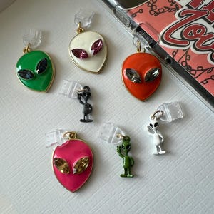 May include: A collection of colorful alien-themed phone charms. The charms feature various colors, including green, white, orange, and pink, with gold accents and gem-like eyes. Small alien figurines are also included.