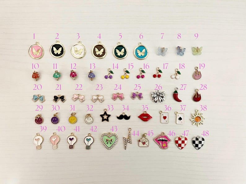 May include: A collection of 48 colorful enamel charms in various shapes, including butterflies, mushrooms, cherries, bows, stars, mustaches, lips, playing cards, hearts, and lightbulbs. Each charm has a gold-tone metal border.