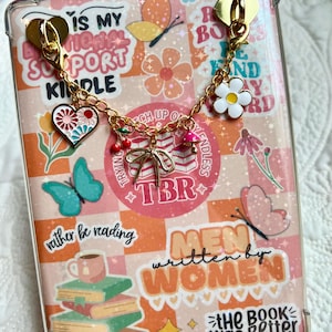 May include: A clear Kindle case with a decorative charm. The case features a colorful design with text and illustrations of books, butterflies, and flowers. The charm includes a heart, cherries, and a daisy. The text includes "IS MY MENTAL SUPPORT KINDLE" and "MEN written by WOMEN".