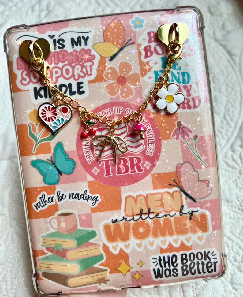May include: A clear Kindle case with a decorative charm. The case features a colorful design with text and illustrations of books, butterflies, and flowers. The charm includes a heart, cherries, and a daisy. The text includes "IS MY MENTAL SUPPORT KINDLE" and "MEN written by WOMEN".