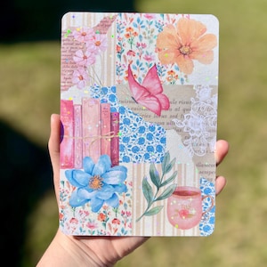 May include: A pink and blue floral patterned notebook with a butterfly, books, a cup of tea, and a sprig of green leaves. The notebook has a white lace border and glitter accents.