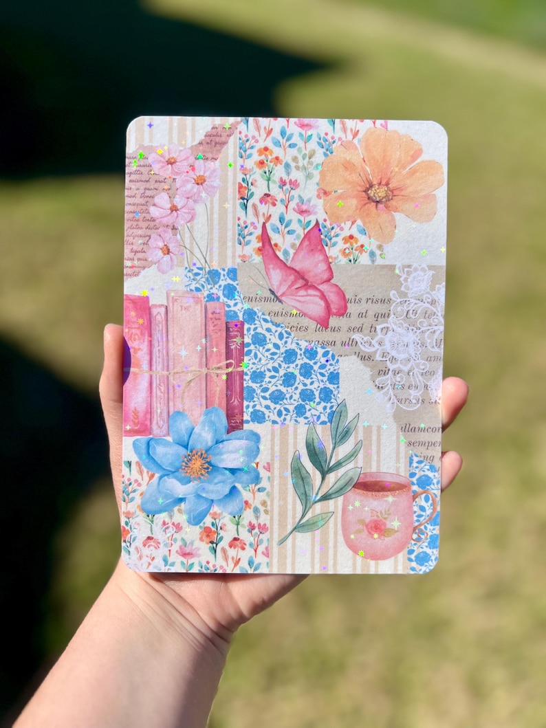 May include: A pink and blue floral patterned notebook with a butterfly, books, a cup of tea, and a sprig of green leaves. The notebook has a white lace border and glitter accents.
