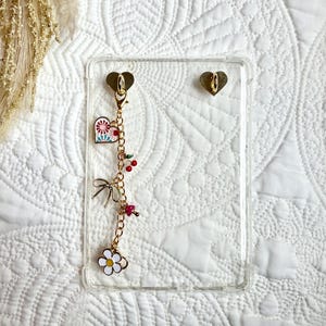 May include: A clear phone case with gold heart-shaped attachments. A gold chain with charms, including a heart, cherries, a bow, a mushroom, and a daisy, hangs from the case. The case is on a white quilted surface.