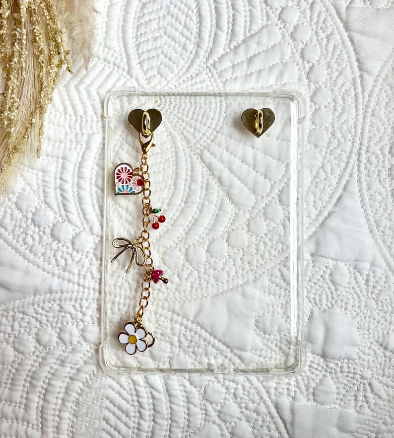 May include: A clear phone case with gold heart-shaped attachments. A gold chain with charms, including a heart, cherries, a bow, a mushroom, and a daisy, hangs from the case. The case is on a white quilted surface.