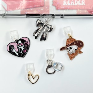 May include: Five different charms for a phone case. The charms are a silver bow, a pink heart with a masked figure holding a knife, a skull wearing a cowboy hat, a gold heart, and silver handcuffs.