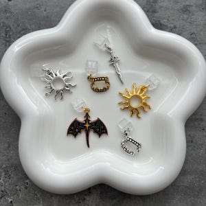 May include: A collection of small, decorative charms in various shapes, including suns, fangs, a sword, and a dragon. The charms are silver, gold, and black, and are displayed on a white, cloud-shaped dish.