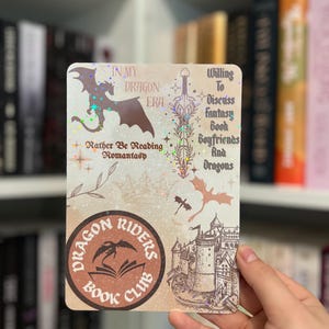 May include: A rectangular sticker with a fantasy theme, featuring a dragon silhouette, a castle illustration, and the text "Dragon Riders Book Club." Additional text includes "In My Dragon Era" and "Rather Be Reading Romantasy."
