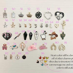 May include: A collection of metal charms in various shapes, including stars, crowns, hearts, and a cactus. The charms are in silver, gold, black, and other colors. Text on the image says "Heart color will be chosen based off of other charms picked and vibes due to inventory!"