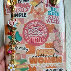 May include: A clear phone case with a colorful design featuring the text "IS MY EMOTIONAL SUPPORT KINDLE" and "MEN written by WOMEN". The case has a gold charm with a heart, flowers, and cherries.