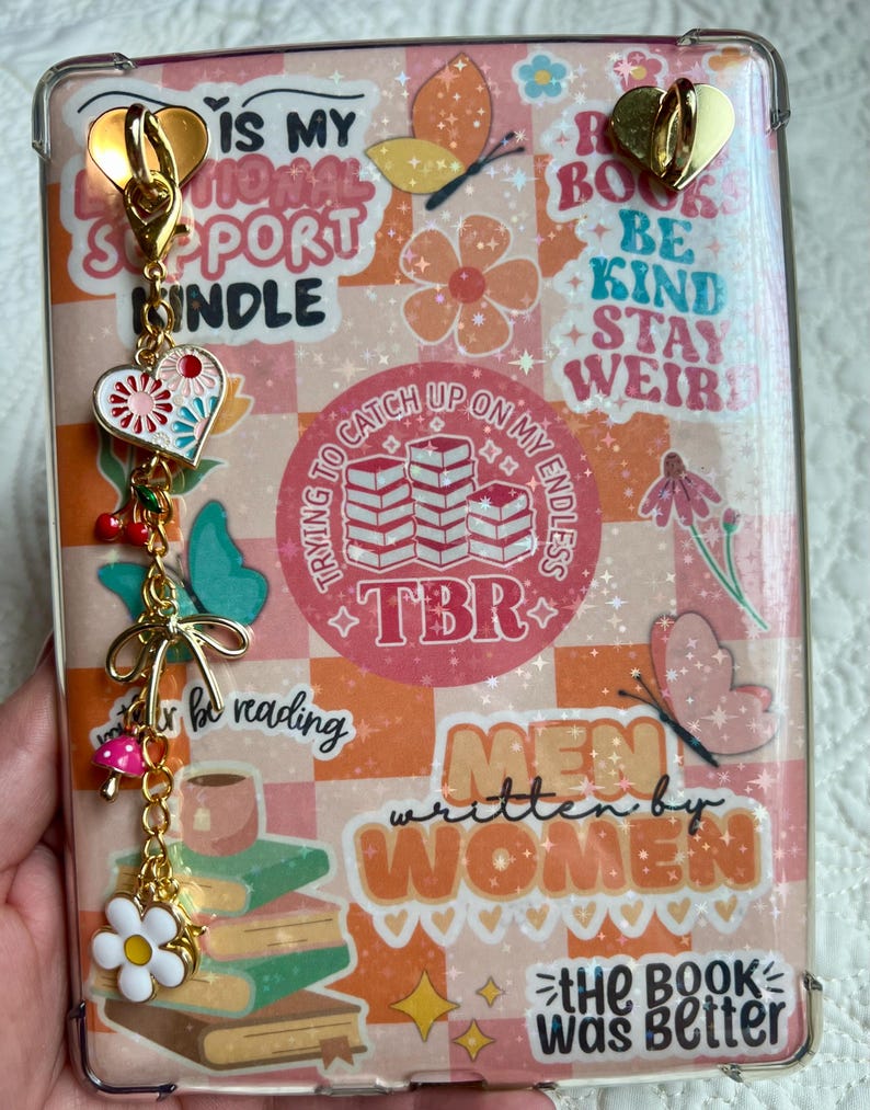 May include: A clear phone case with a colorful design featuring the text "IS MY EMOTIONAL SUPPORT KINDLE" and "MEN written by WOMEN". The case has a gold charm with a heart, flowers, and cherries.