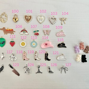 May include: A collection of small, colorful charms in various shapes, including a crescent moon, a padlock, hearts, a bow, a dachshund, a rainbow, cherries, a butterfly, a cowboy hat, an alien, a strawberry, a daisy, a cowboy boot, a knife, and gummy bears.