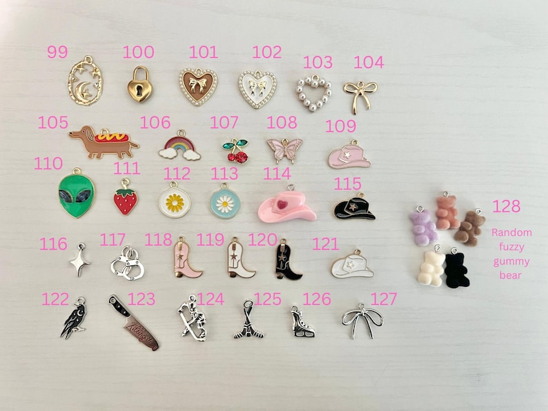 May include: A collection of small, colorful charms in various shapes, including a crescent moon, a padlock, hearts, a bow, a dachshund, a rainbow, cherries, a butterfly, a cowboy hat, an alien, a strawberry, a daisy, a cowboy boot, a knife, and gummy bears.