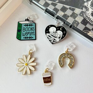 Puede incluir: Cinco charms diferentes para el teléfono con clips de plástico transparente. Los charms son: un libro azul con el texto "BOOK WORM", un corazón blanco y negro con dos esqueletos abrazándose, una margarita blanca, una herradura dorada con pedrería y una taza de café blanca con el texto "coffee".