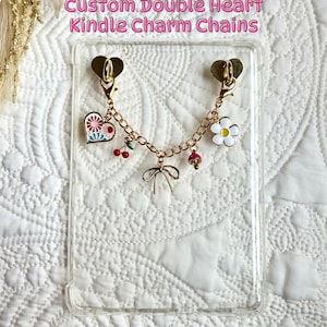 Könnte beinhalten: Eine transparente Kindle-Hülle mit einer goldfarbenen Doppelherz-Charmkette. Die Kette hat verschiedene Charms, darunter ein Herz mit floralem Design, eine weiße Blume, Kirschen und eine Schleife. Der Text "Custom Double Heart Kindle Charm Chains" wird oben angezeigt.