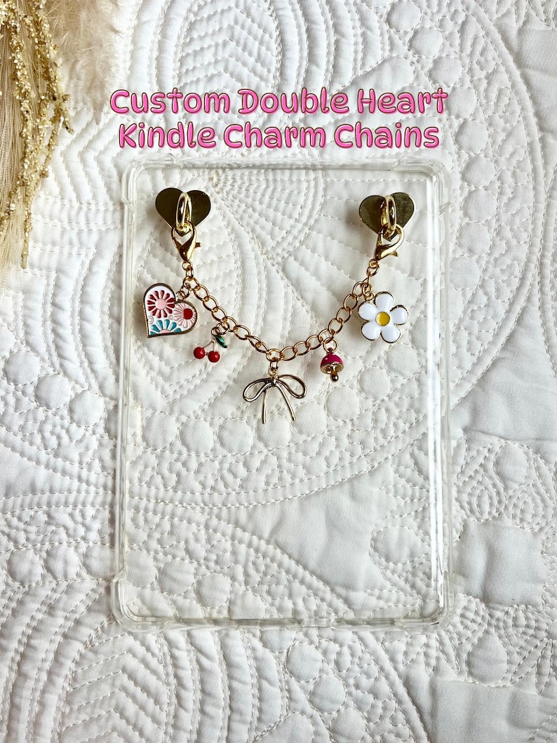 May include: A clear Kindle case with a gold-colored double heart charm chain. The chain features various charms, including a heart with a floral design, a white flower, cherries, and a bow. The text "Custom Double Heart Kindle Charm Chains" is displayed at the top.