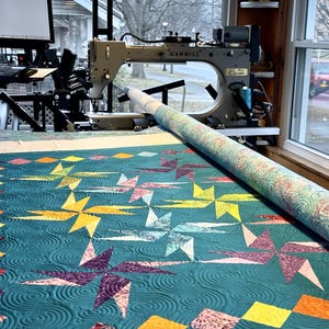 May include: A teal quilt with a starburst pattern is being quilted on a long-arm quilting machine. The machine is a Gammill Classic model. The quilt is being quilted with a swirling pattern.