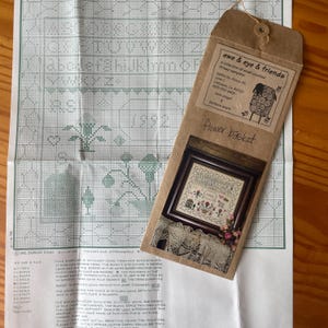 May include: A brown paper envelope with a white label that reads "ewe & eye & friends" and "flower basket". The envelope contains a cross-stitch pattern with a floral design and the text "1992".