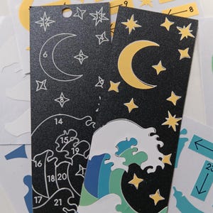 May include: Two black bookmarks with celestial designs. One features a crescent moon and stars, the other a wave design. Additional stickers with arrows and numbers are also visible.