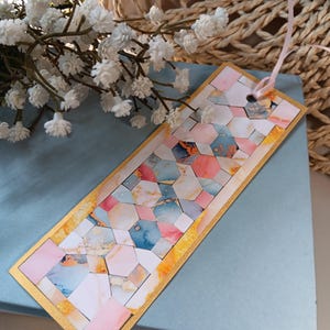May include: A rectangular bookmark with a gold border and a colorful geometric design. The design features pink, blue, and gold hexagonal shapes. A pink ribbon is attached at the top. The bookmark rests on a blue surface.