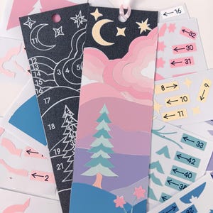 May include: A layered paper bookmark featuring a night sky with a crescent moon and stars above pink clouds, a pine tree, and blue hills with pink flowers. A second black bookmark with white line art and numbers is visible alongside sticker sheets.